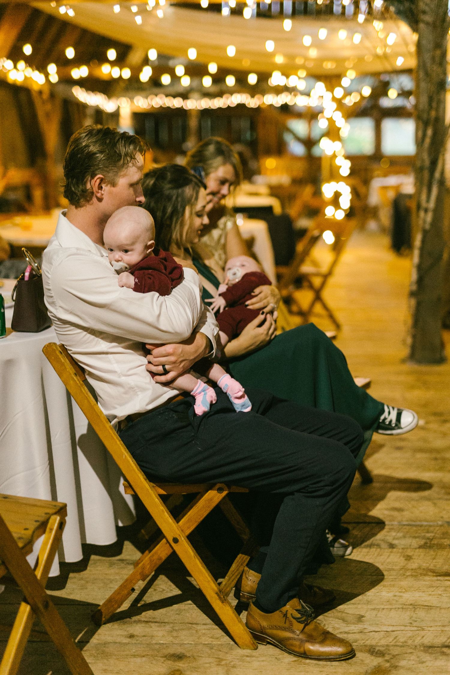 Parent holding baby while seated on wooden chairs at an evening event with string lights overhead.