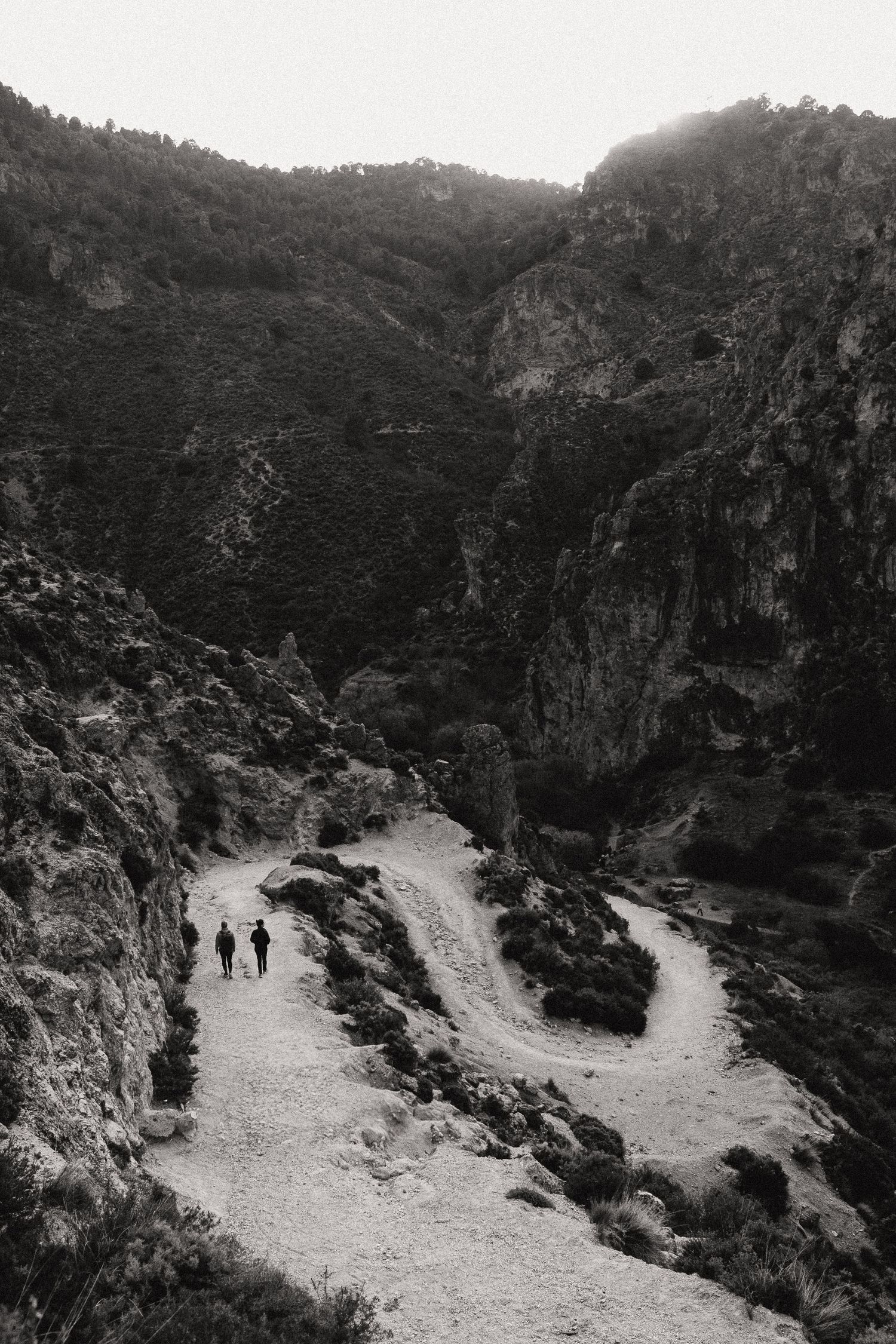 Two distant hikers walk along a winding mountain path through rugged terrain in black and white.