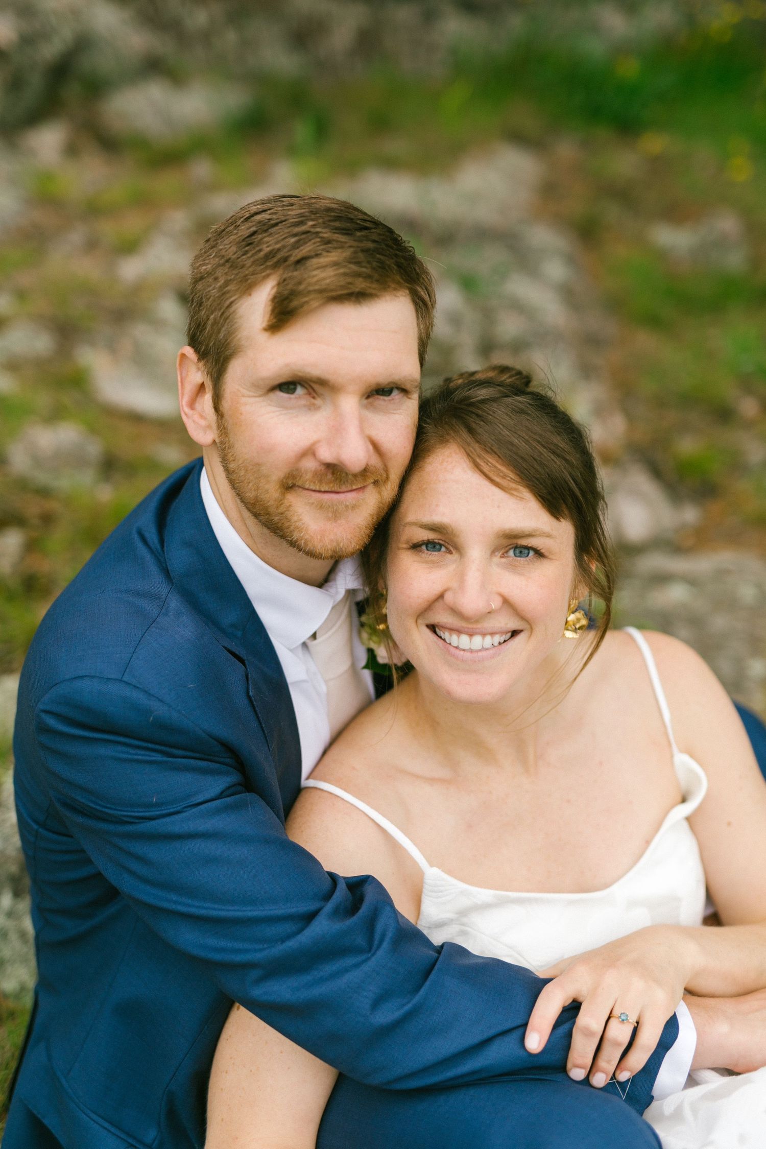 A smiling couple in wedding attire embrace outdoors against a natural backdrop with soft lighting.
