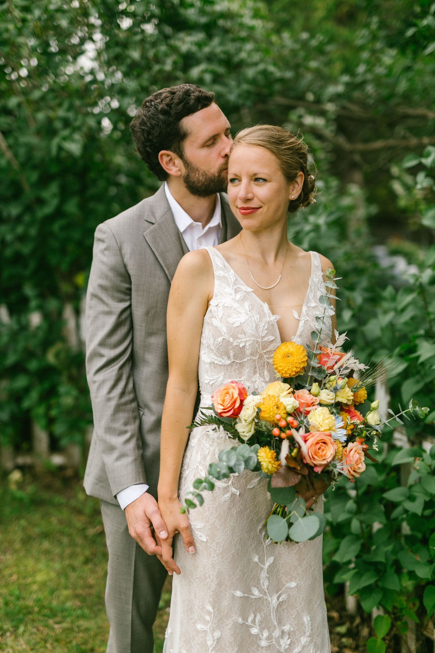 Newlywed couple embraces in garden setting with coral and yellow floral bouquet and gray suit and lace dress.