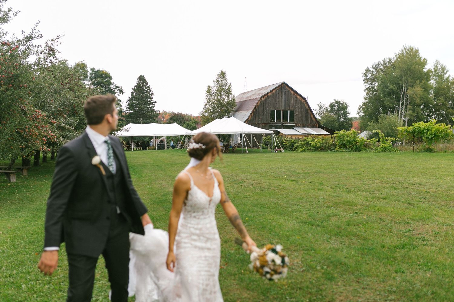 A couple walk across a rustic farm property with a barn and white tent in the background.