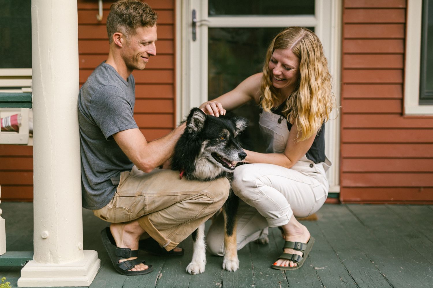 A couple pets their husky dog while sitting on a front porch.