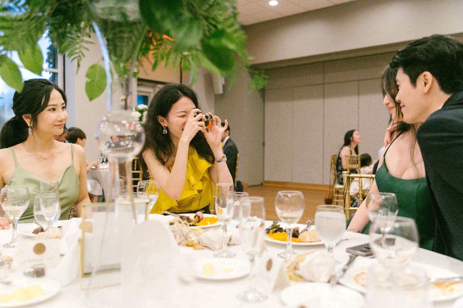 Three guests laughing and taking photos at an elegant wedding reception dinner table with floral decorations.