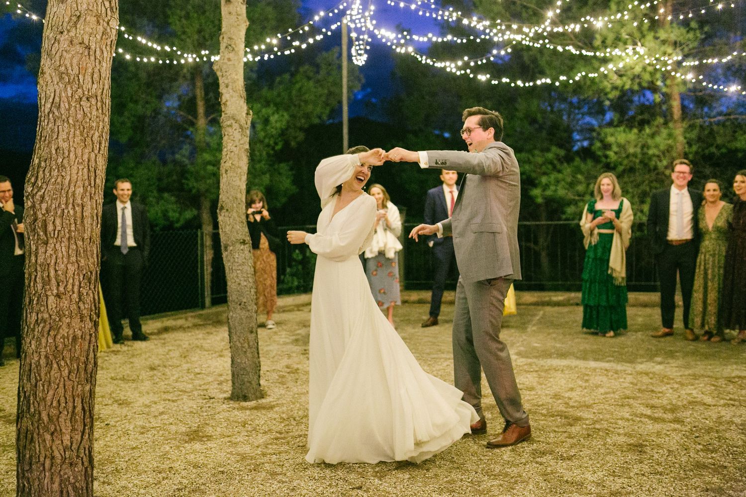 A couple shares their first dance at a romantic outdoor wedding reception under twinkling string lights between trees.