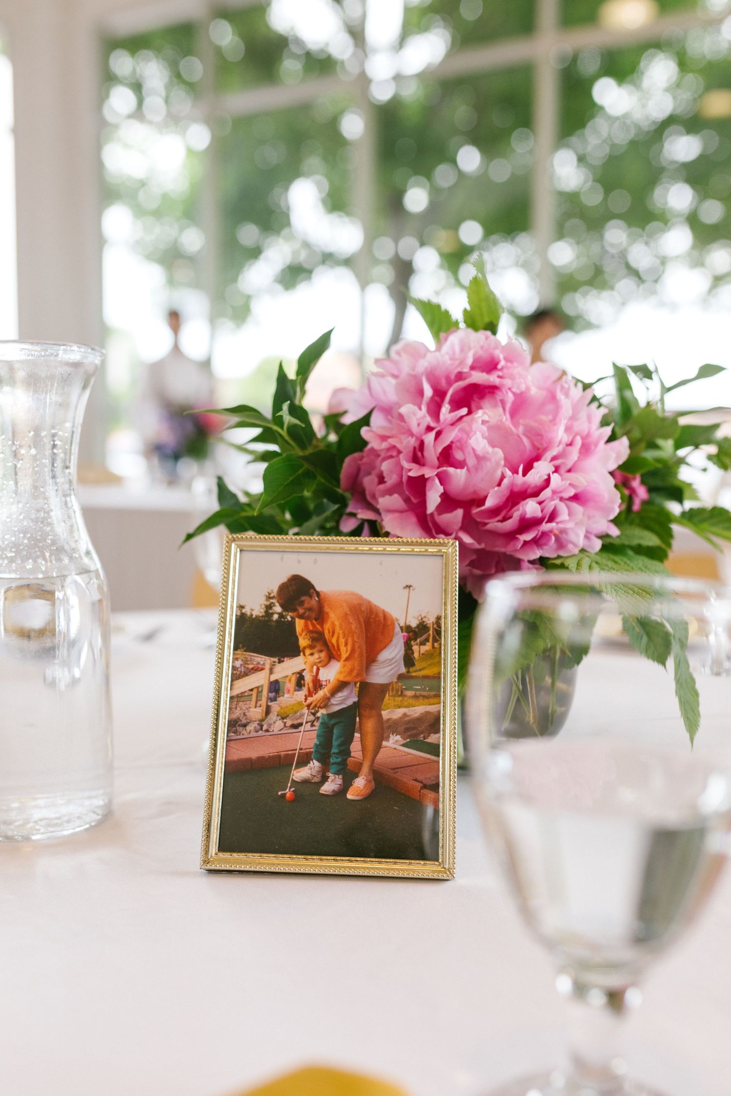 A pink peony flower arrangement next to a framed photograph on a white tablecloth with glassware.