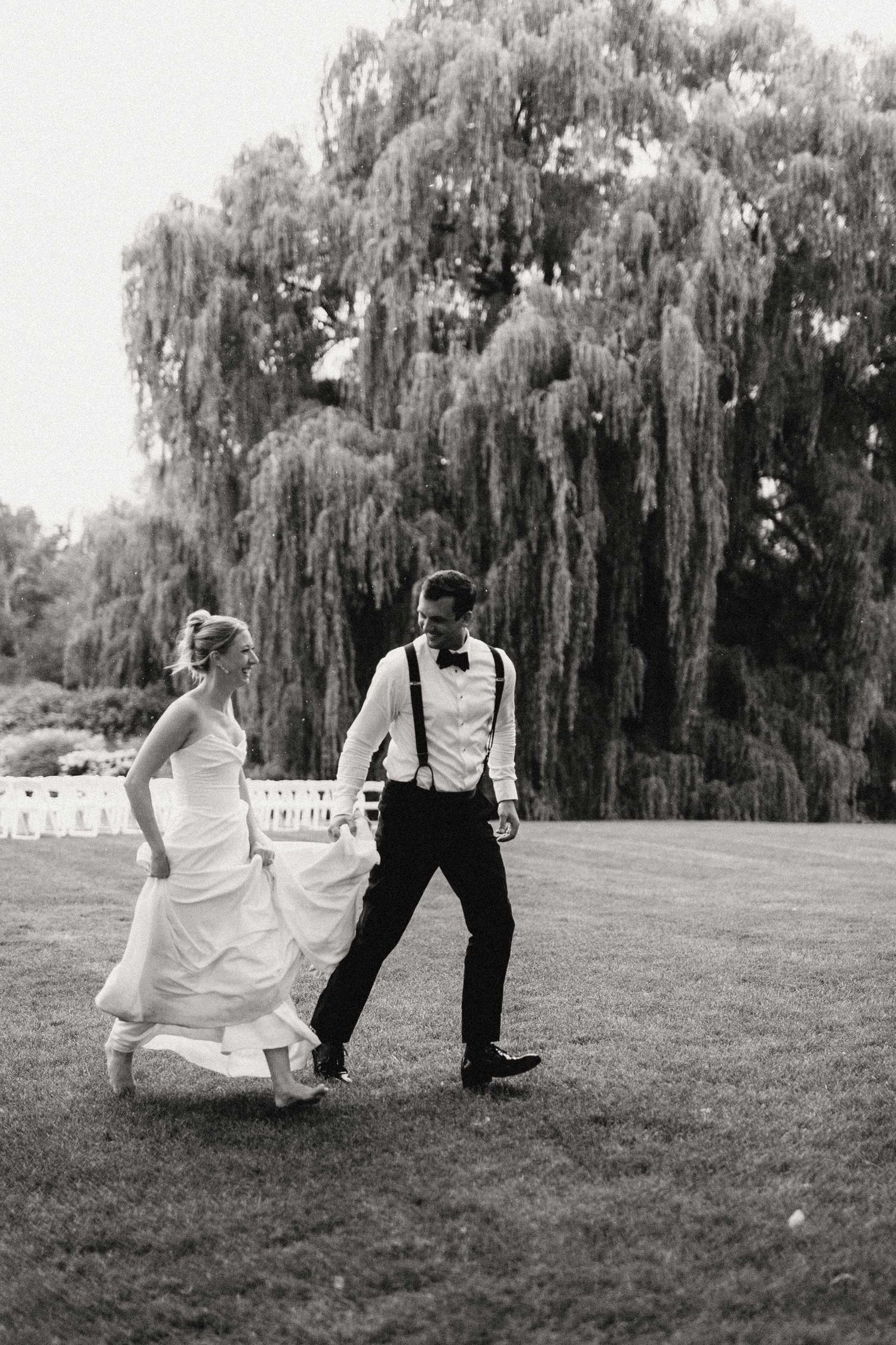 A black and white wedding photo showing a couple dancing playfully on a lawn with a majestic weeping willow tree in background.