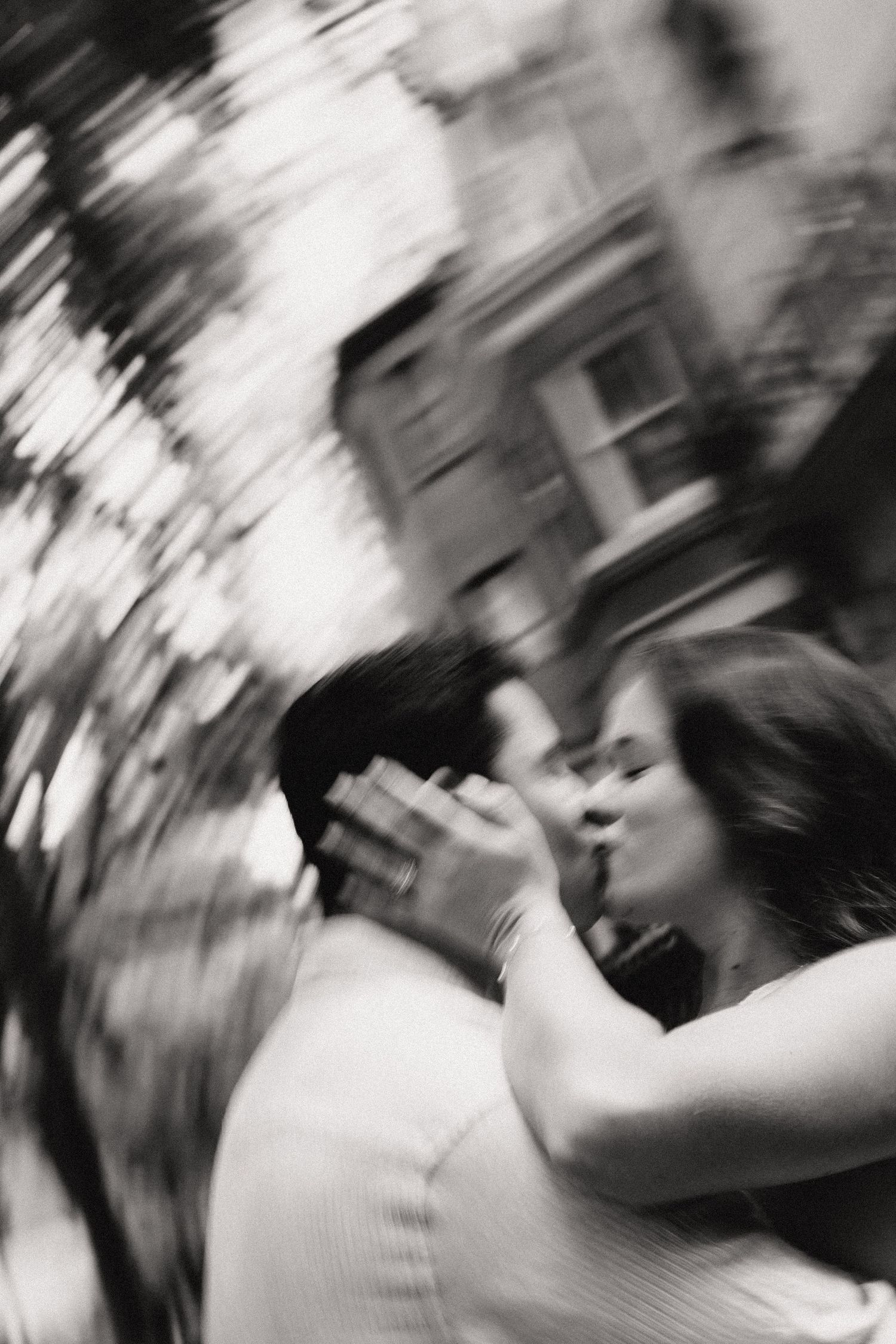 Black and white blurred photo of a couple sharing a romantic kiss outdoors near trees and buildings.