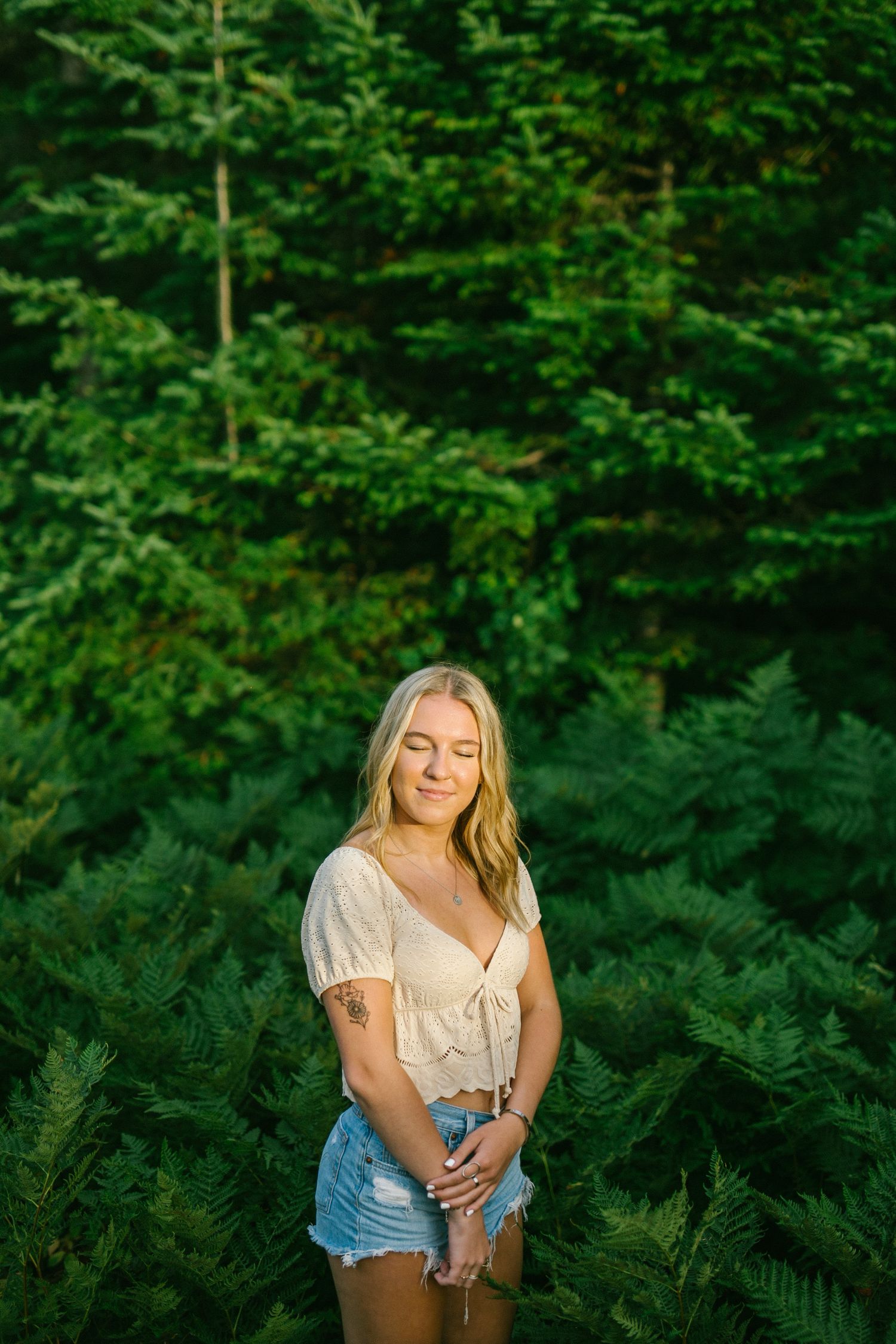 Blonde woman in white top and denim shorts standing among lush green ferns and pine trees in sunlight.