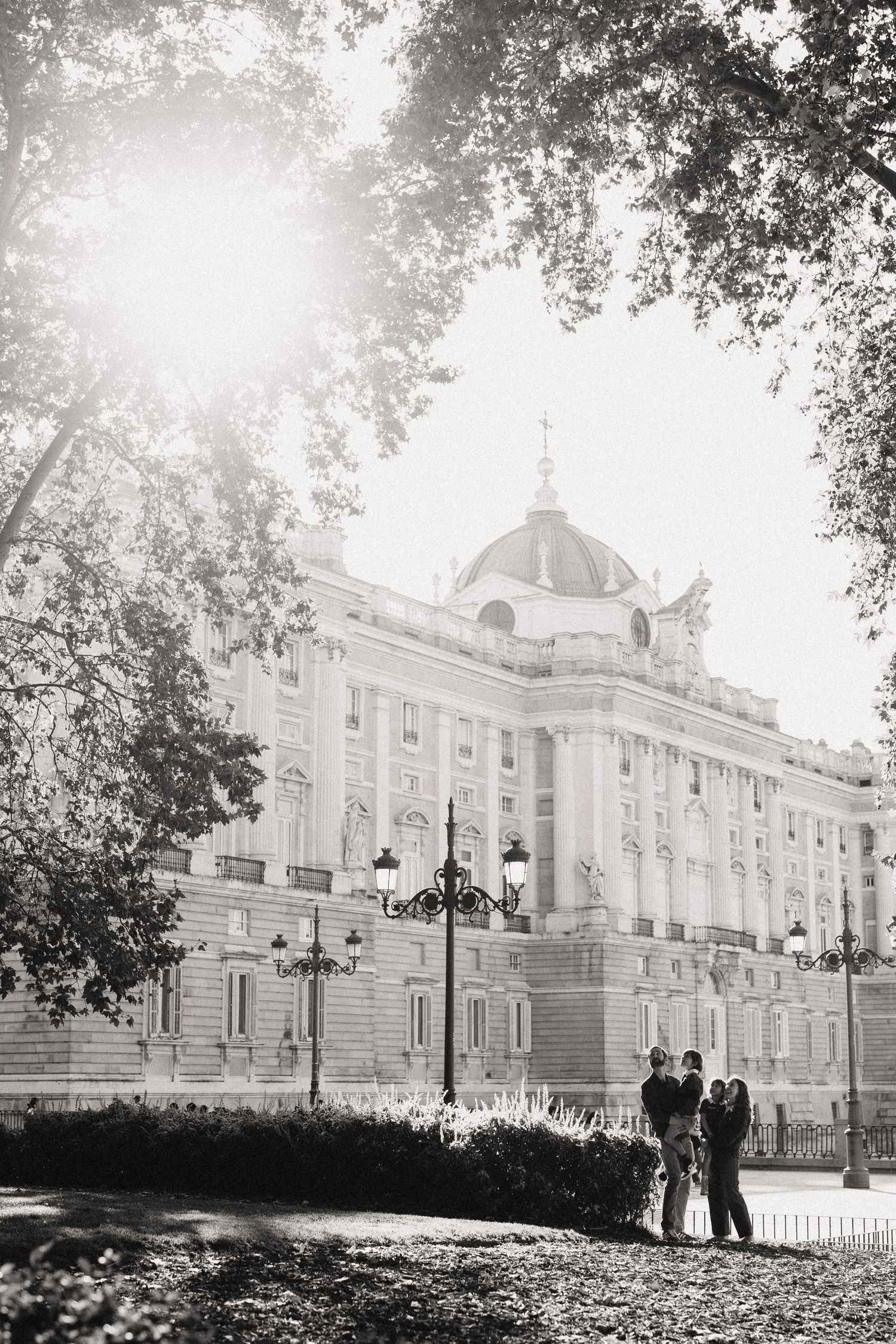 Black and white photo of a couple standing in a park with the Royal Palace of Madrid in the background.