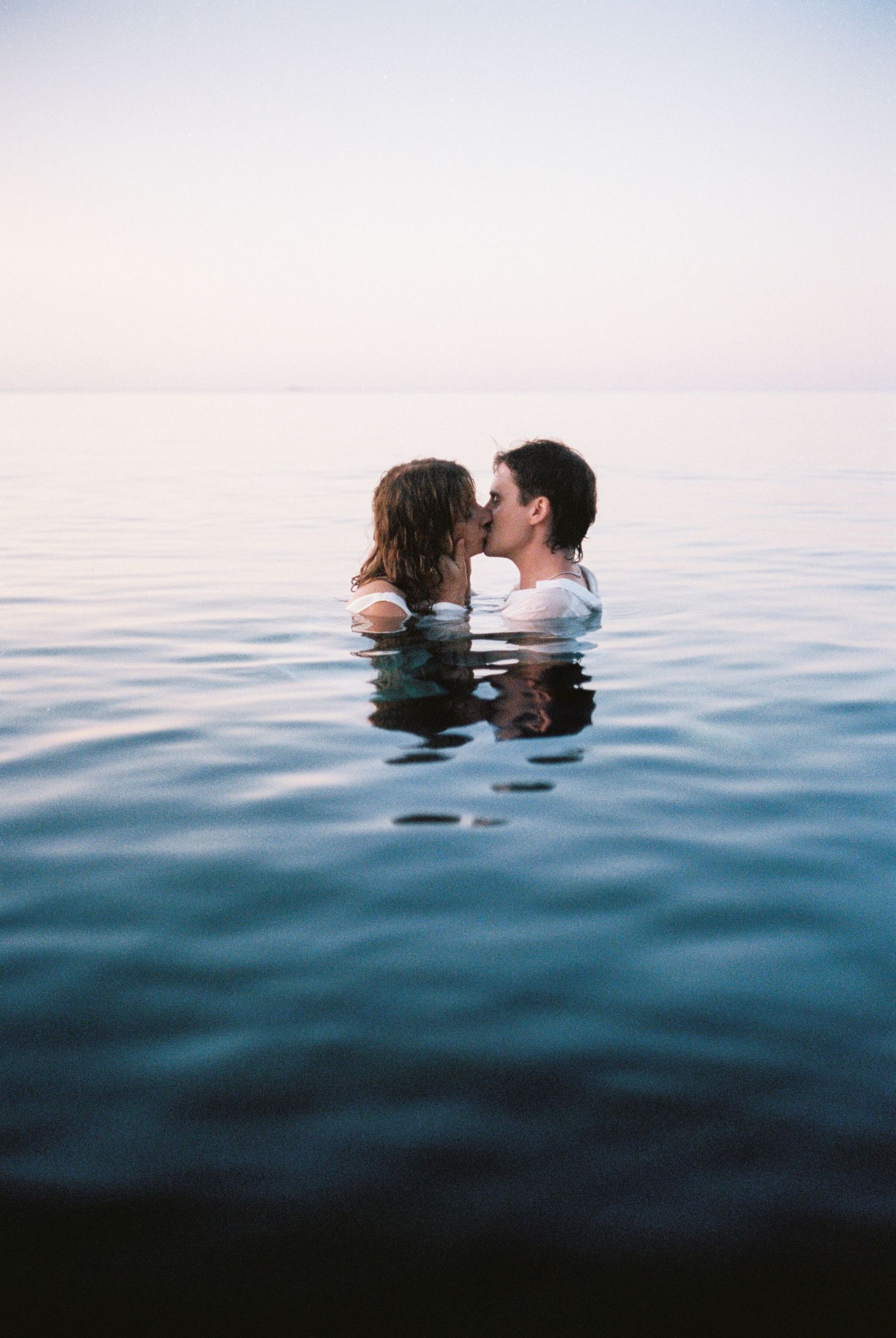 Romantic couple kissing while swimming in calm ocean water at dusk.