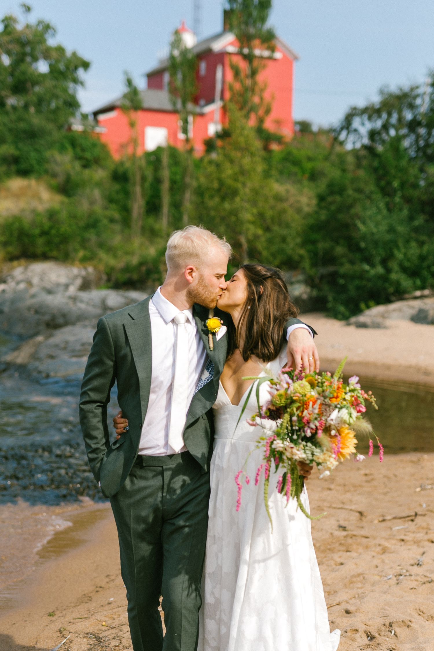 Newlyweds share intimate moments along a sandy beach with a red lighthouse in the distance.