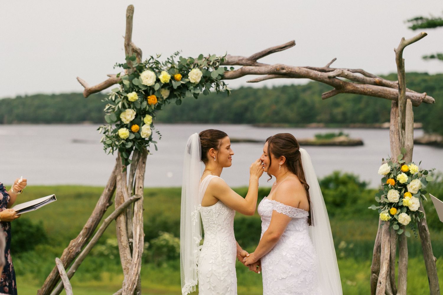 Two brides in white dresses exchange vows under a rustic driftwood arch with floral decor by a scenic waterfront.