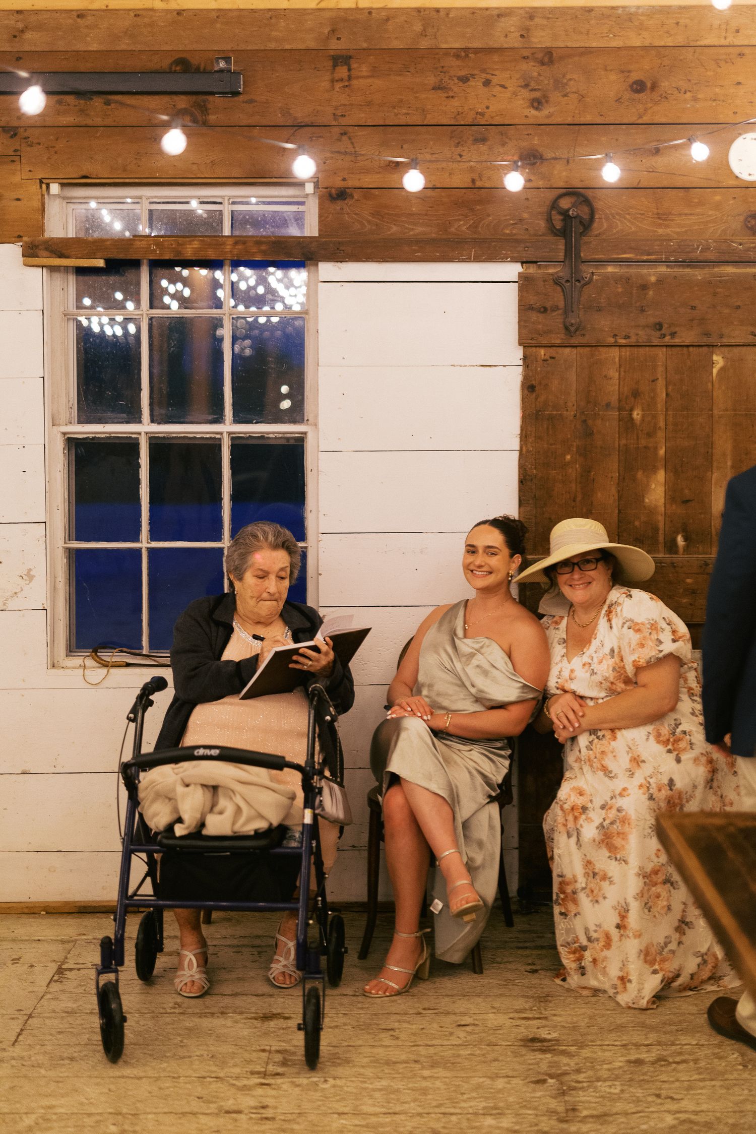 Three women seated in a rustic barn venue with string lights, one using a walker, at a wedding reception.