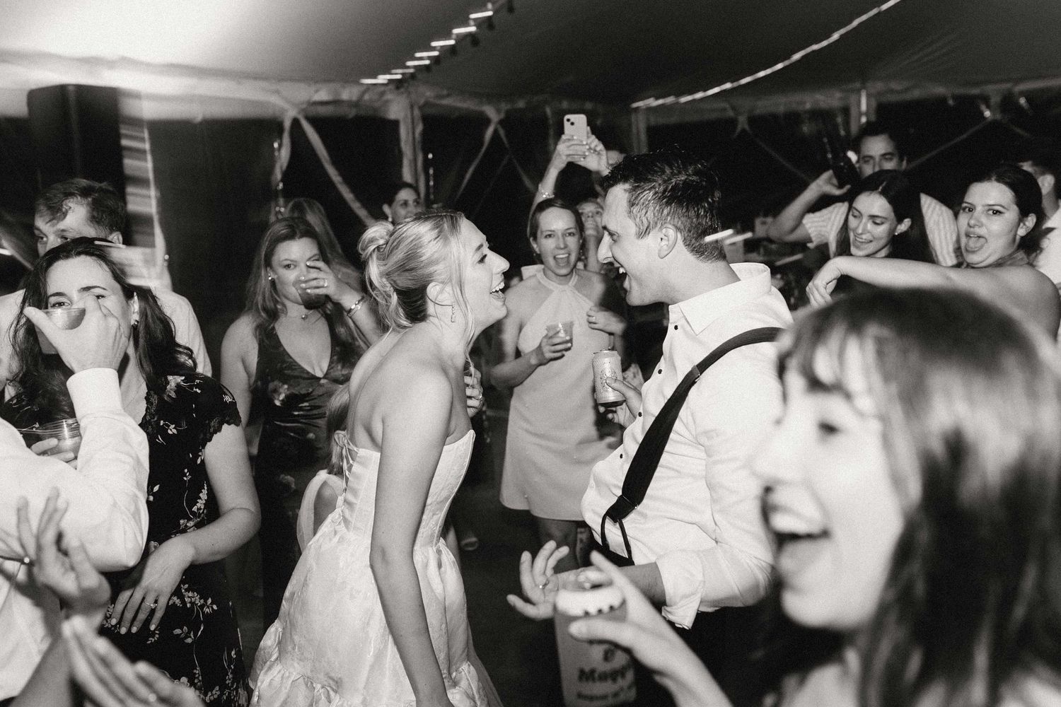 A black and white photo of a a bride and groom surrounded on a dance floor with people dancing and celebrating the reception.
