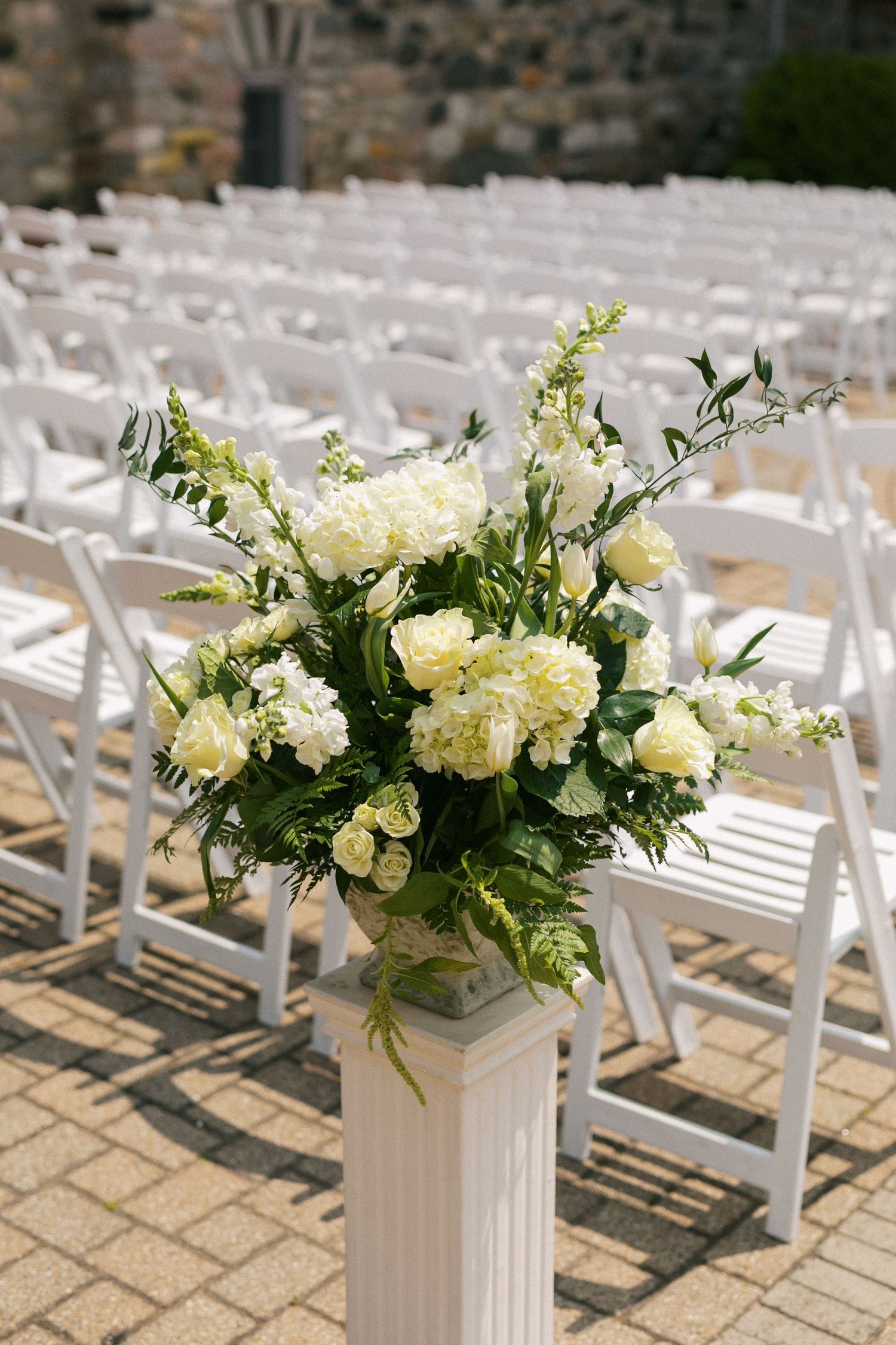 White floral arrangements in columnar vases line the aisle of an outdoor wedding ceremony venue.
