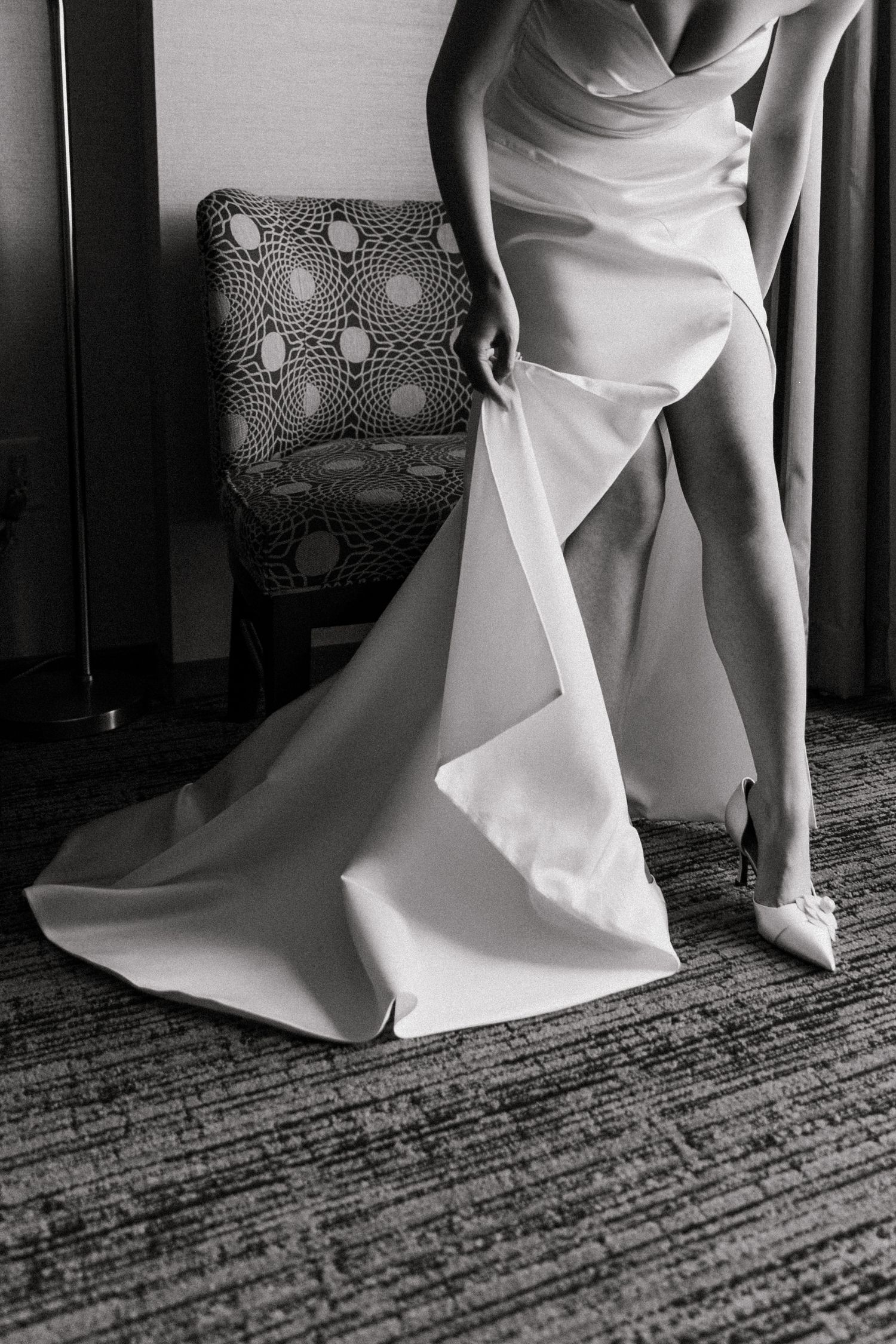Black and white photo of a bride in a sleek wedding gown with a flowing train, wearing heels in an elegant room.