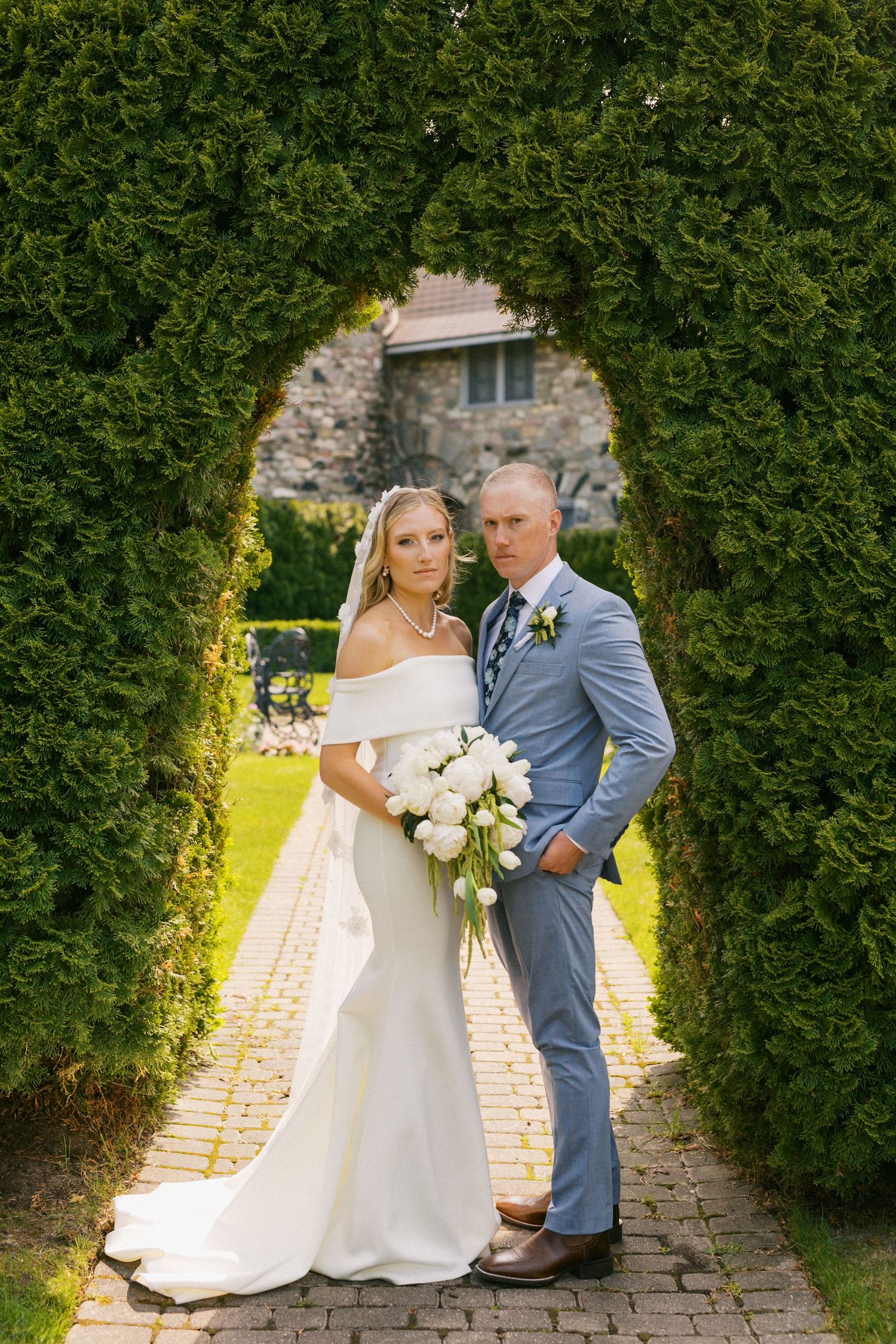 Elegant wedding portrait in a manicured garden pathway with lush green hedges and brick walkway.