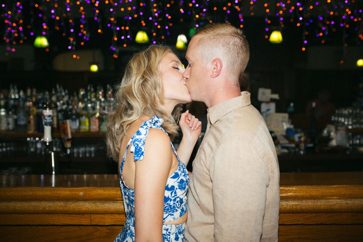A couple shares a romantic kiss at a bar with neon lights and colorful decorations in the background.