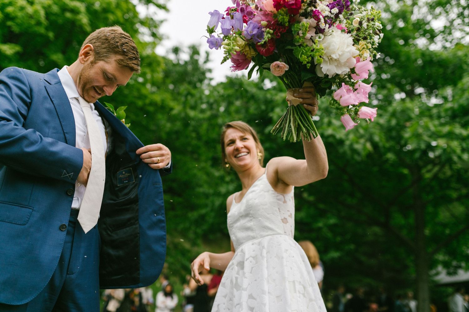 A joyful wedding celebration outdoors featuring a beautiful white dress and colorful raised bouquet.