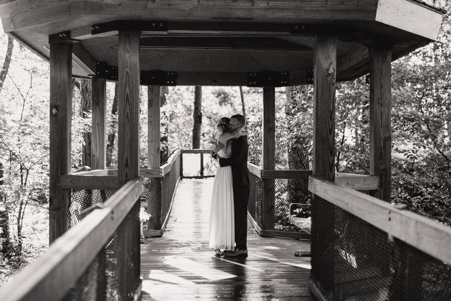 Black and white photo of couple standing on wooden boardwalk surrounded by trees.