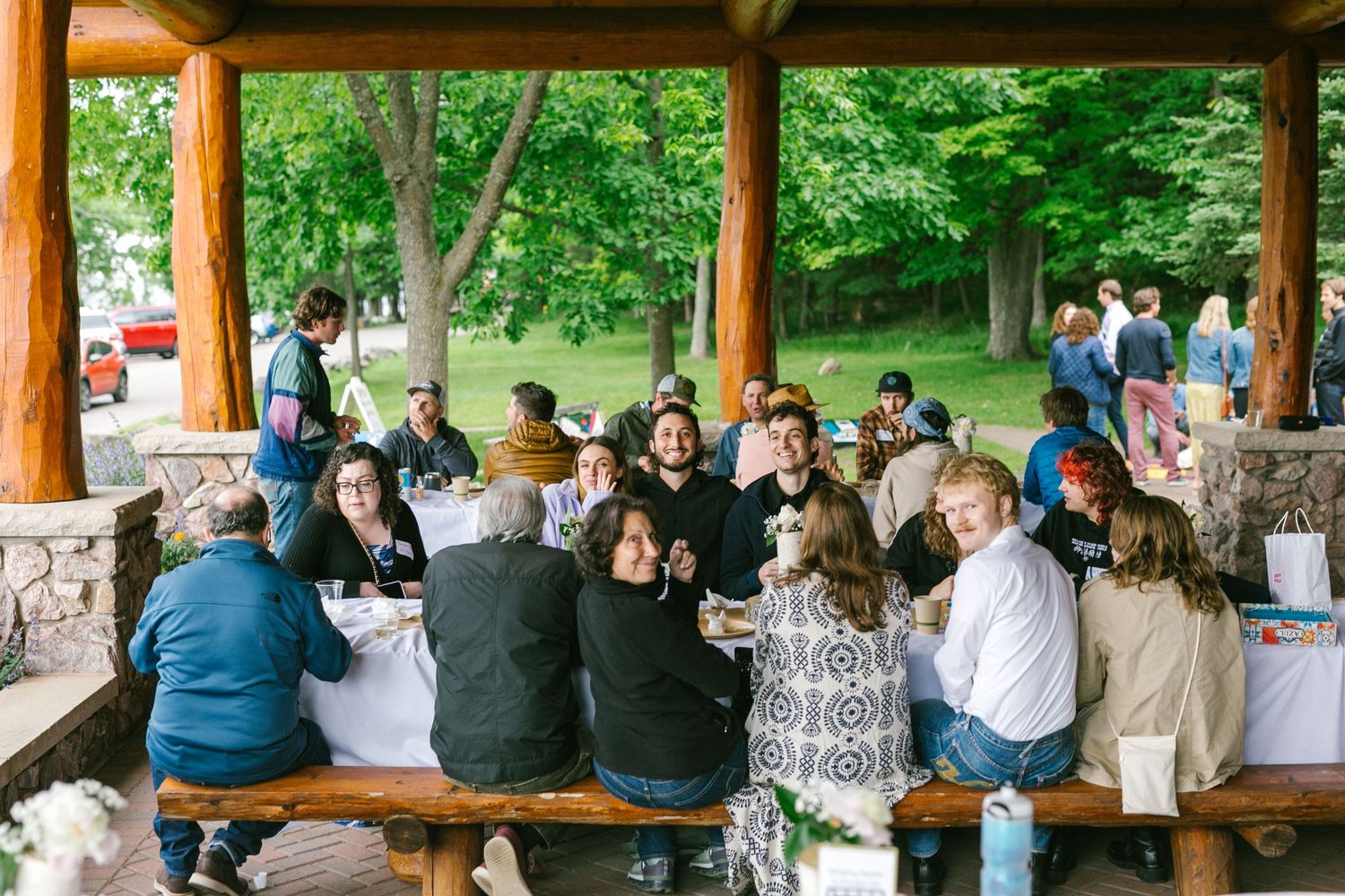People gather under a wooden pavilion roof during an outdoor event in a lush green park setting.