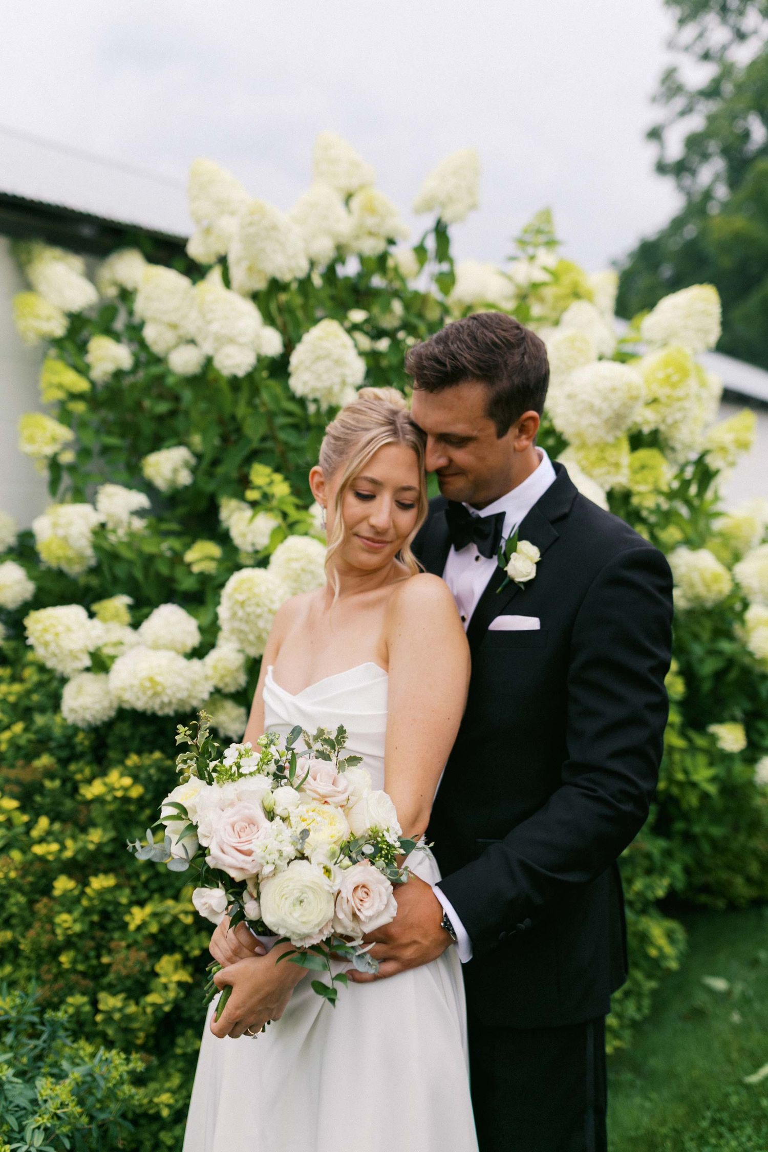 A romantic wedding couple embraces among blooming white hydrangea bushes in a garden setting.