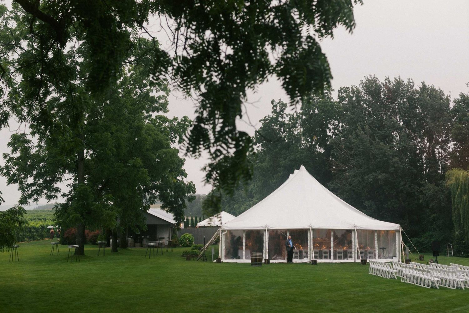 A scenic outdoor wedding venue with white tents and chairs set up on a lush green lawn surrounded by mature trees.
