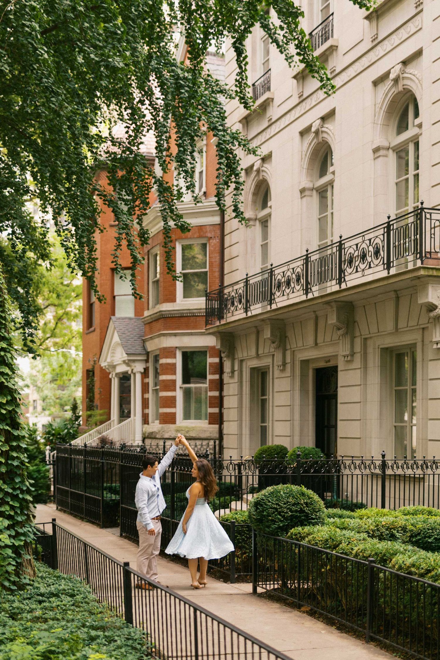 Couple twirling joyfully on a charming urban sidewalk lined with lush greenery and architecture.