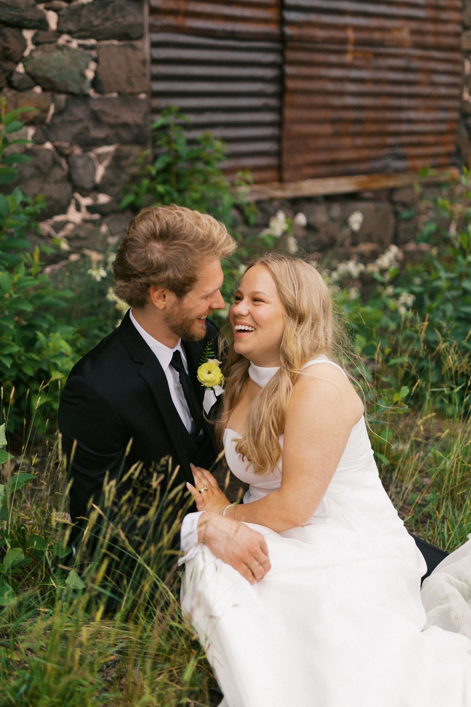Laughing bride and groom sitting in lush green grass near rustic stone wall on wedding day.