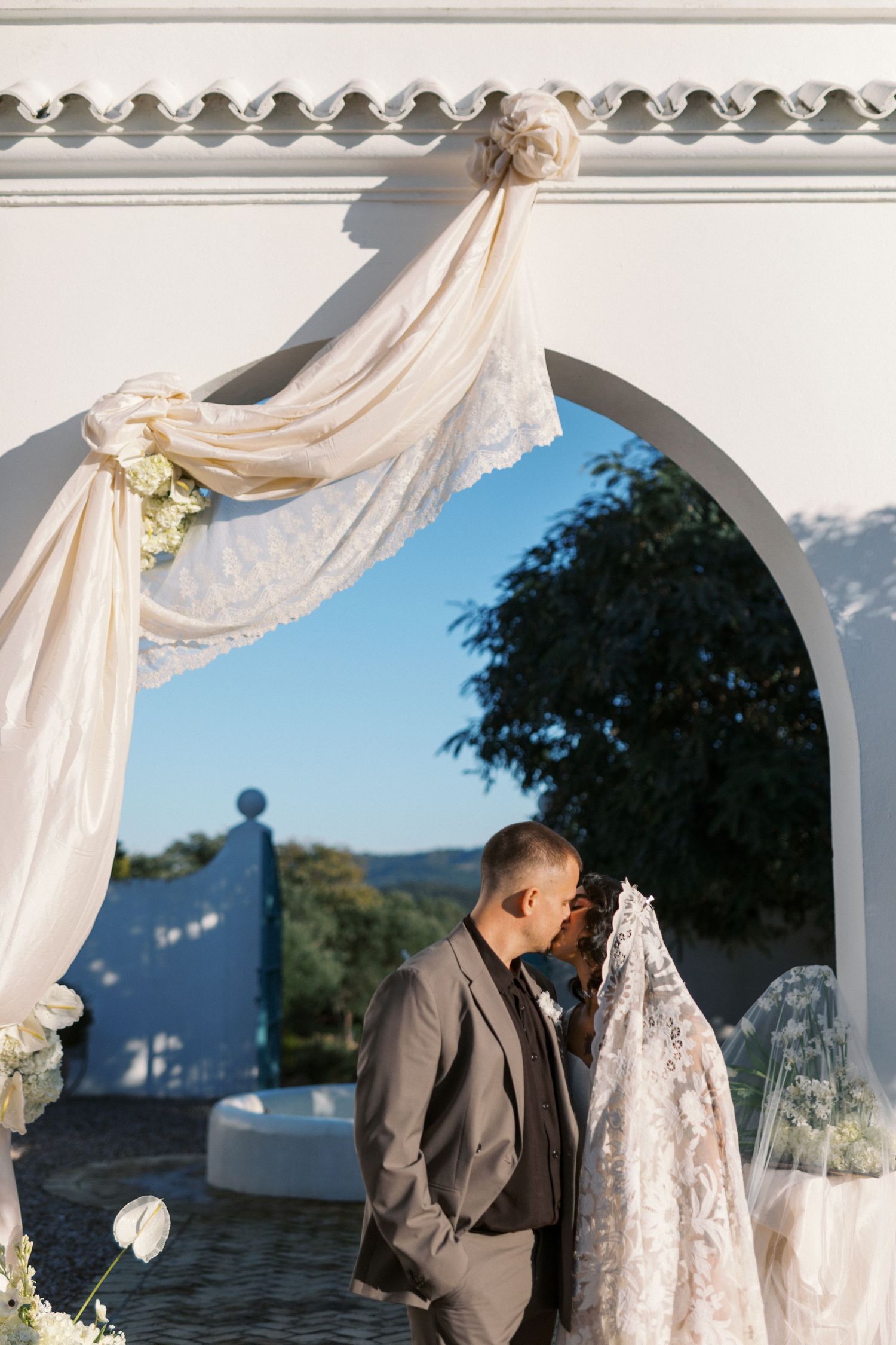 Newlyweds share a kiss under elegant white arch adorned with draped lace fabric at sunny outdoor wedding.