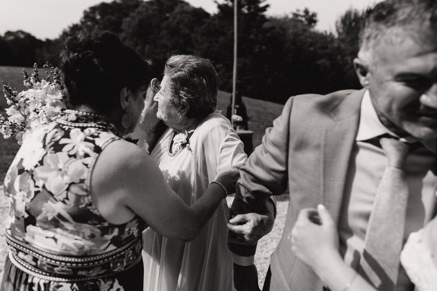 Black and white wedding photo of guests embracing and congratulating the bride outdoors on a sunny day.