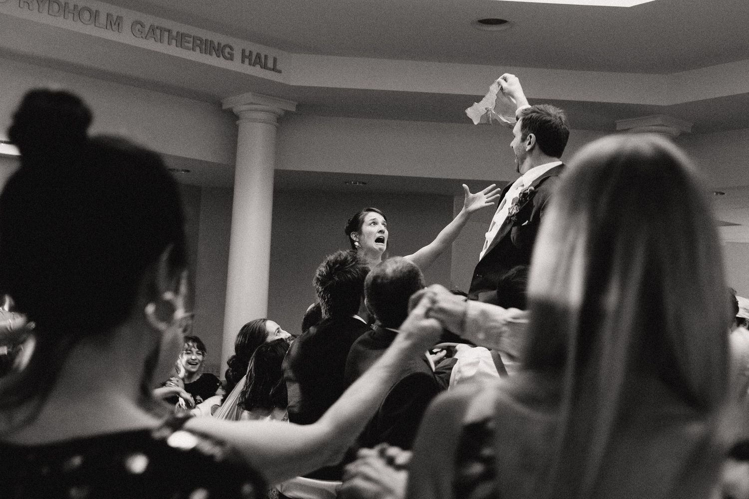 Black and white photo of wedding guests lifting someone during reception celebration.
