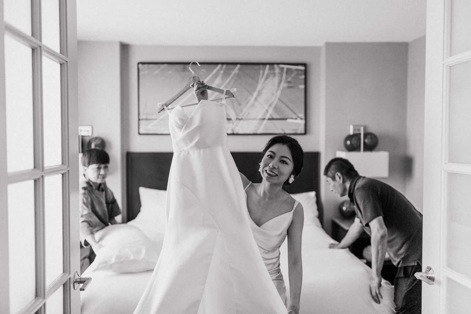 Bride smiling while holding her wedding dress in a hotel room with wedding party preparing in the background.