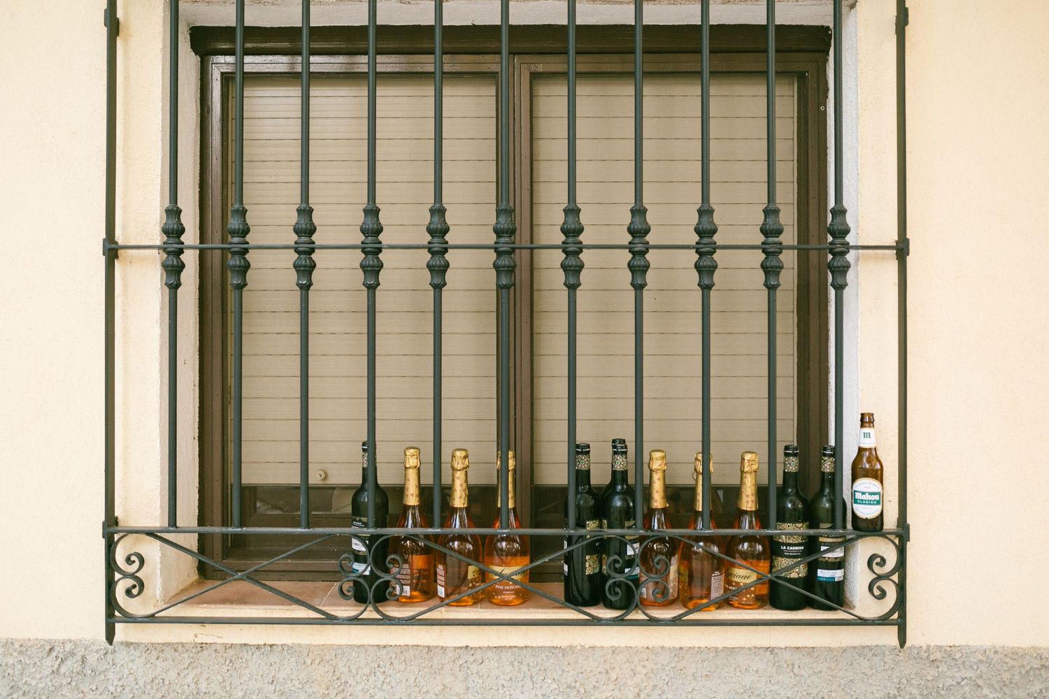 Empty beer and wine bottles displayed on a windowsill behind decorative iron bars in an exterior window.