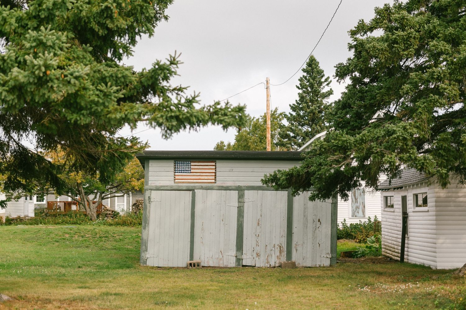 A weathered wooden outhouse structure with white paint stands in a grassy yard surrounded by evergreen trees.