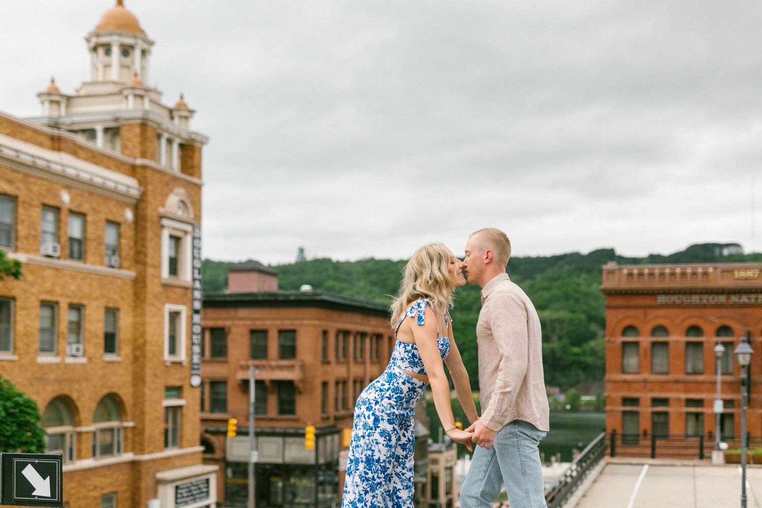 A couple shares a romantic moment on a city rooftop with historic brick buildings and a golden dome cupola in the background.