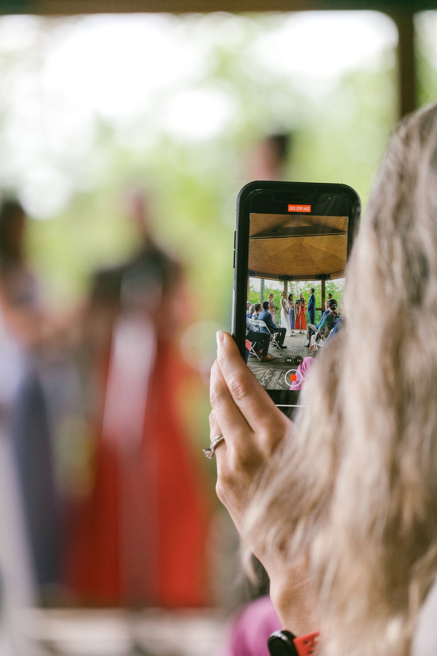 Hand holding phone taking photo of wedding ceremony through smartphone screen.