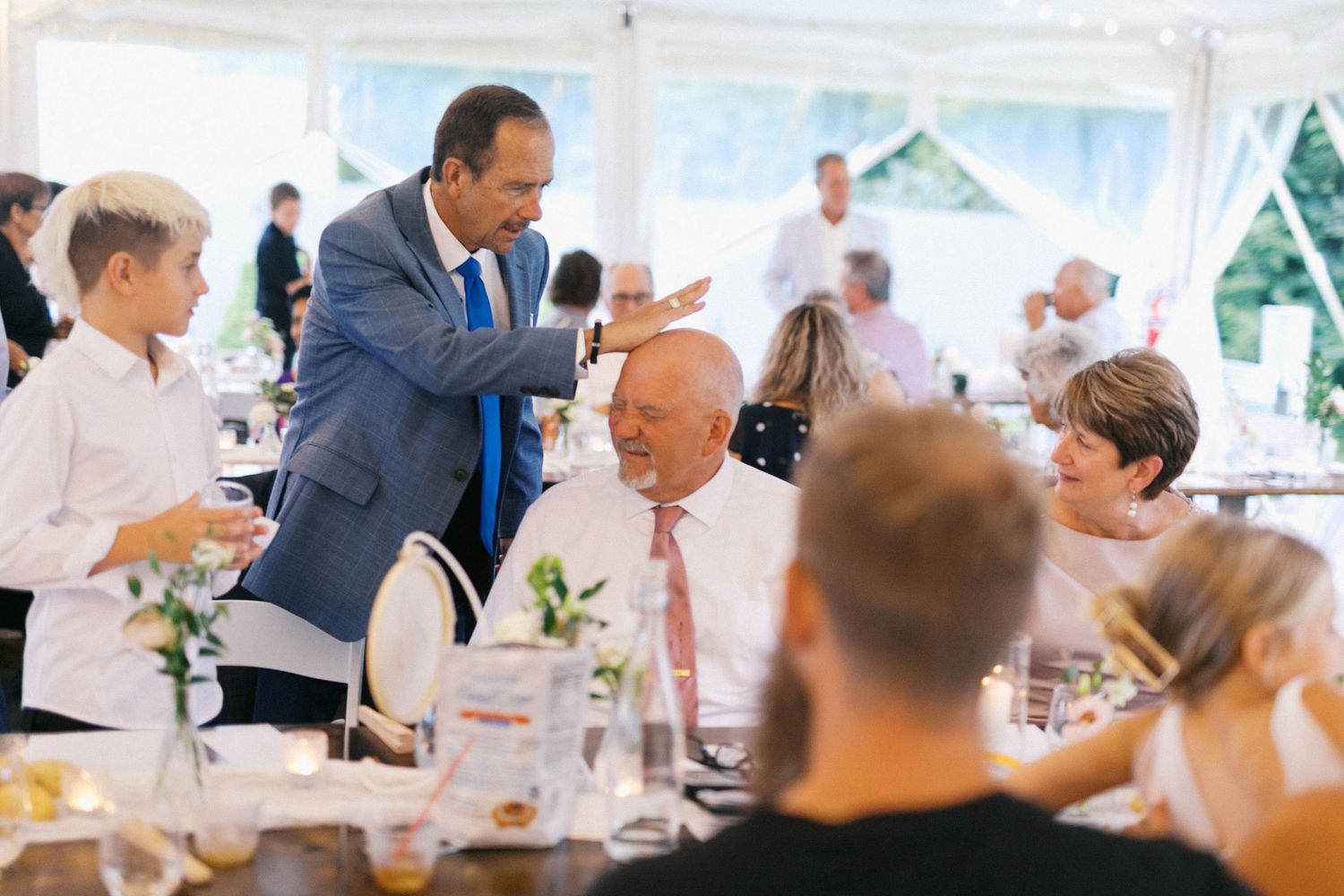 People gathered at a wedding reception under a white tent while someone speaks into a microphone during dinner service.