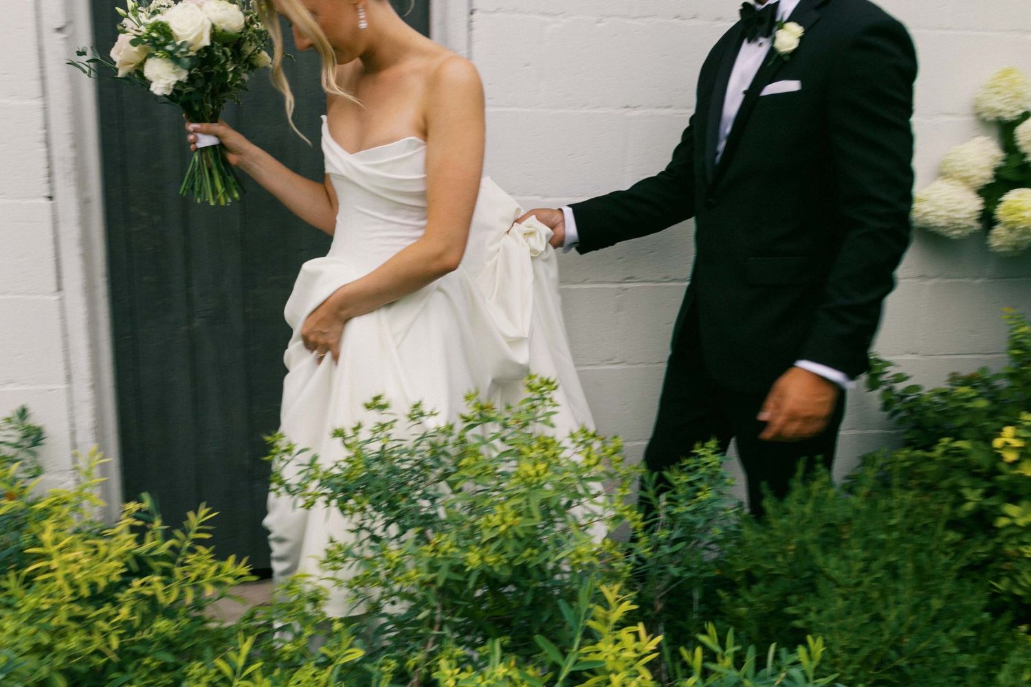 A bride in a white strapless gown and groom in a black tuxedo walk together through lush garden greenery.