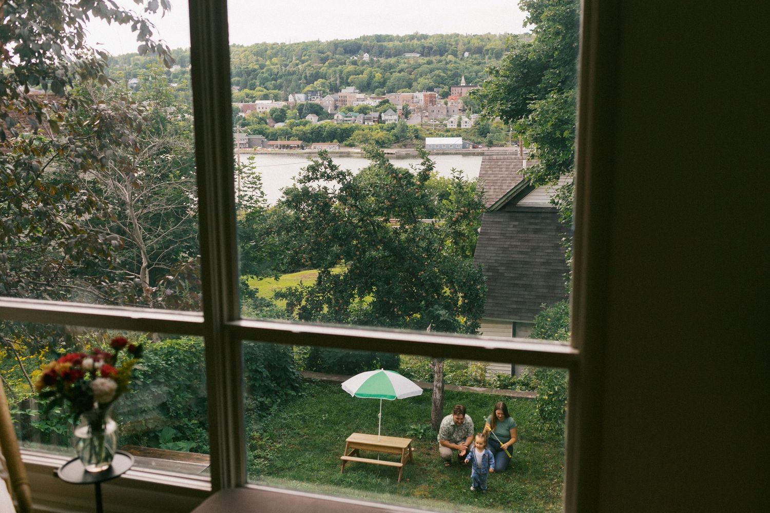 View through window of couple sitting at outdoor picnic table with umbrella overlooking river.