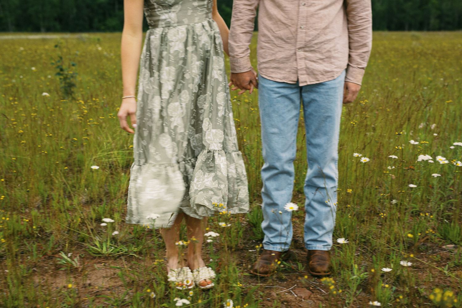 Close-up of couple holding hands in a wildflower field, woman in floral dress and man in jeans.