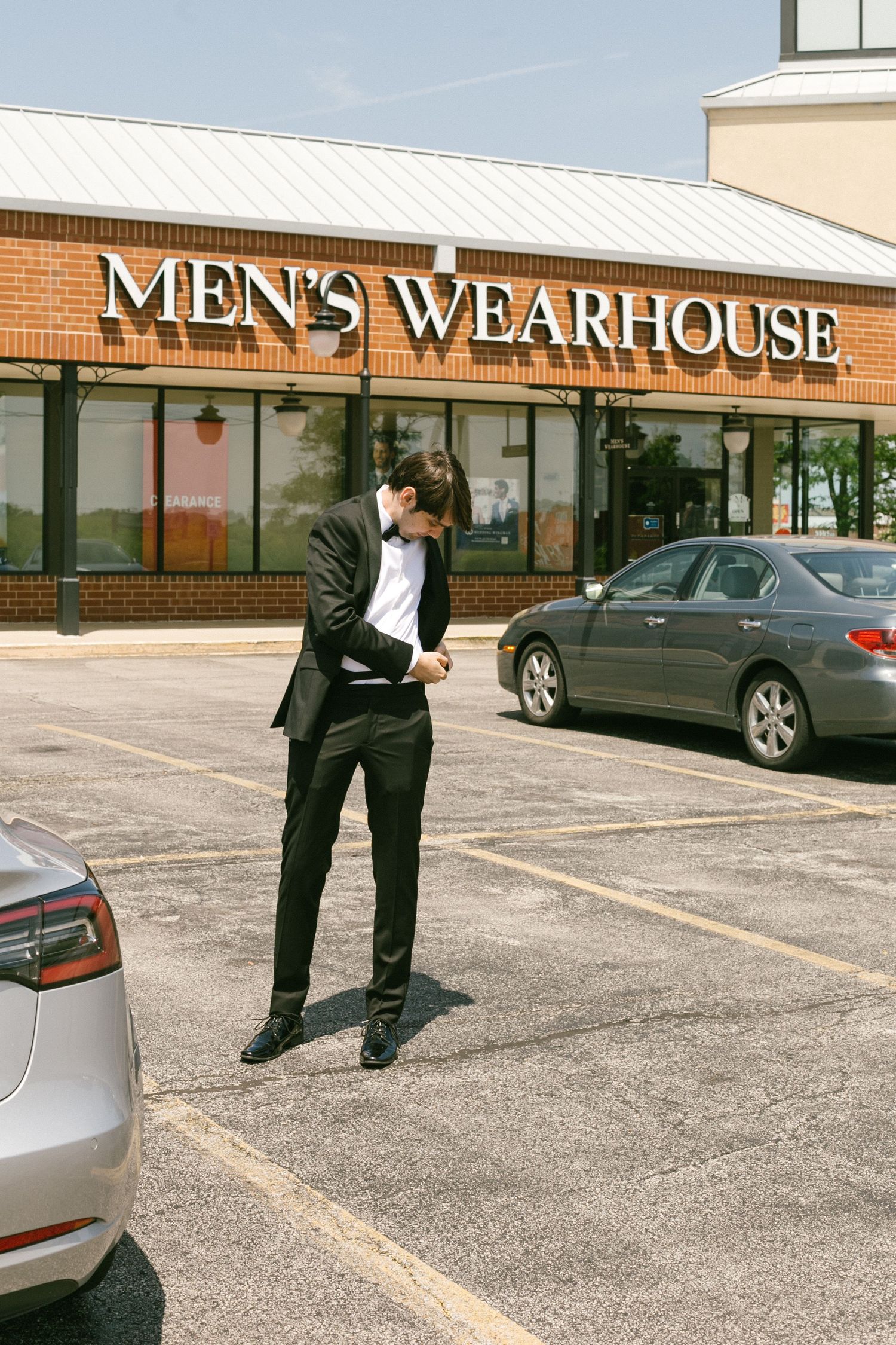 Man in black suit adjusting cufflinks in parking lot outside Men's Wearhouse store.