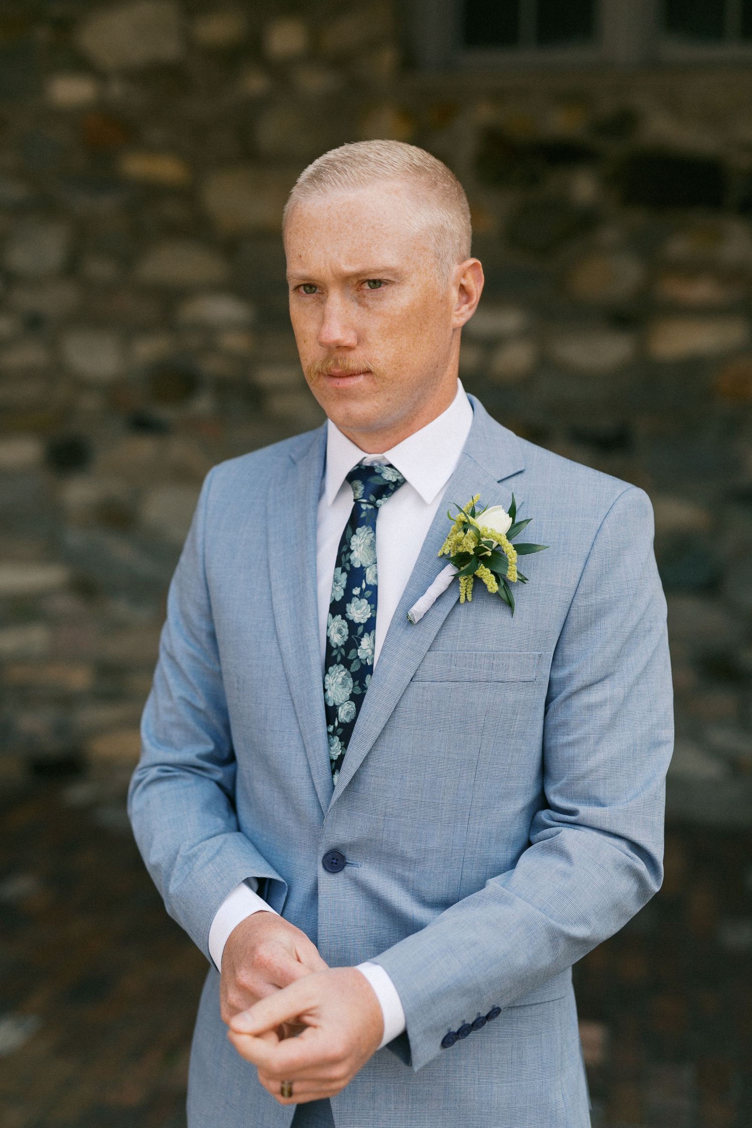 Groom adjusts his tie while wearing a light blue suit with floral tie and boutonniere.