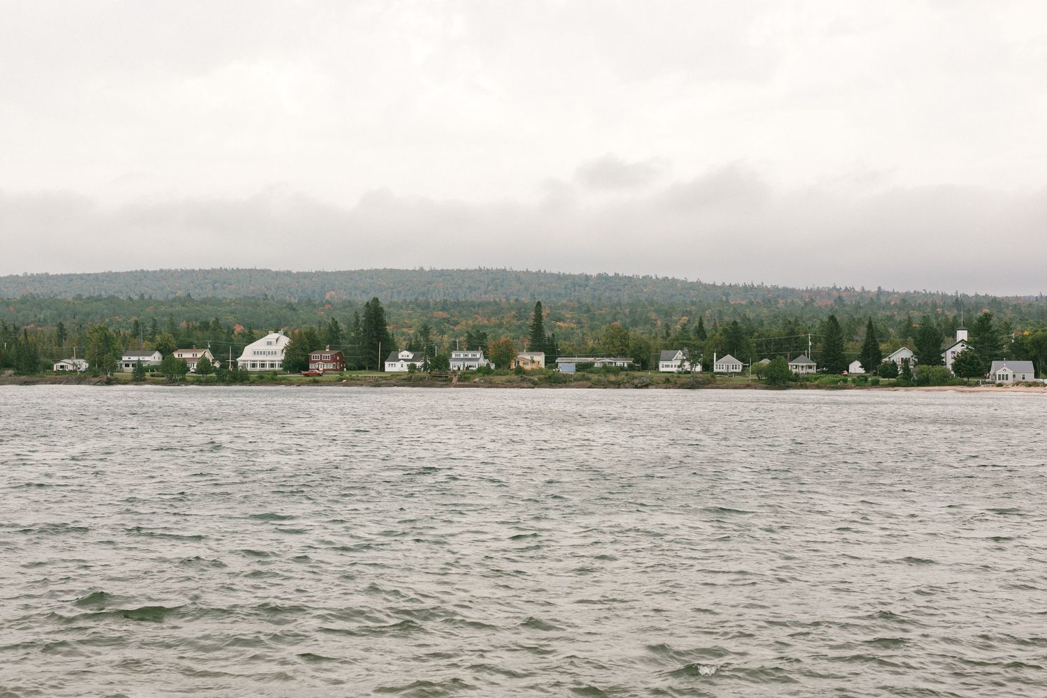 A row of coastal homes along a rugged shoreline with evergreen forests in the background and choppy water in the foreground.
