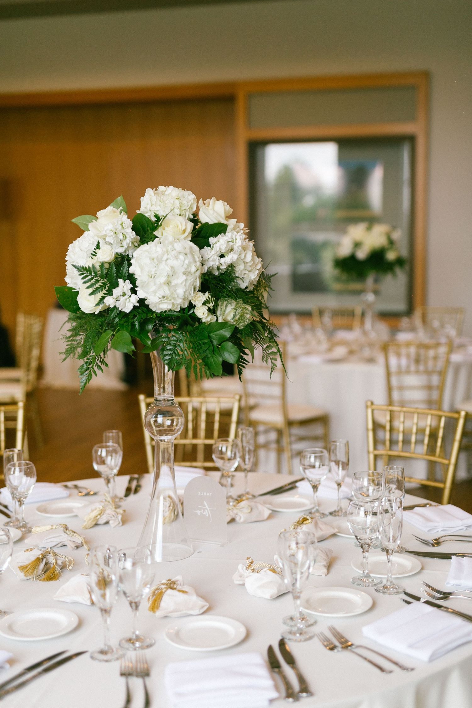 Elegant wedding reception table with tall white hydrangea centerpiece, gold chiavari chairs, and white linens.