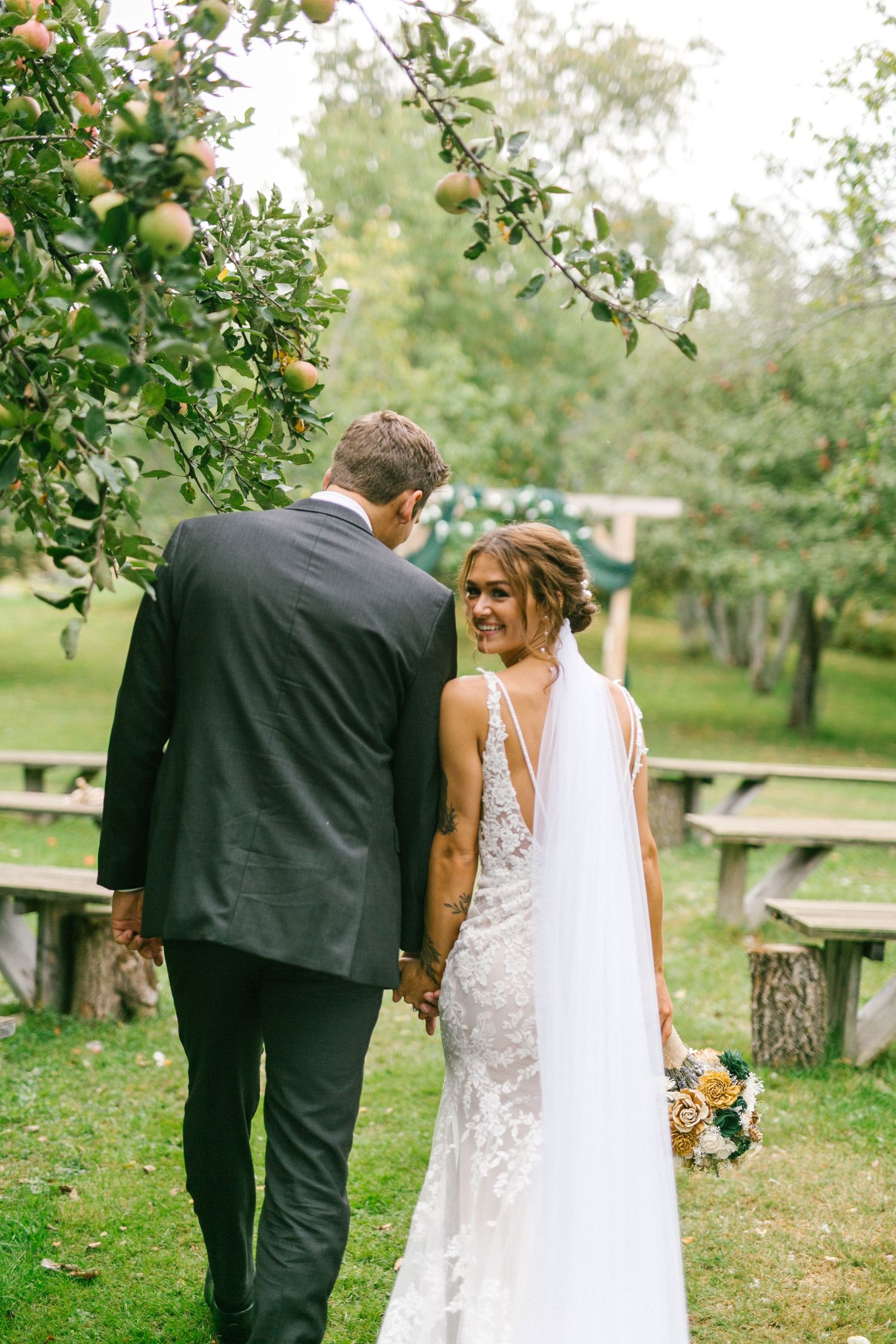 A couple shares a tender moment in an orchard on their wedding day, with apple trees and picnic tables in the background.