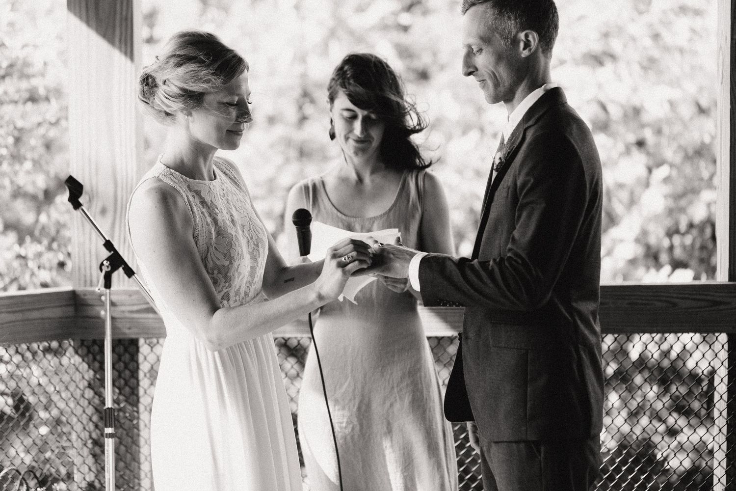 Black and white photo of wedding ceremony with participants exchanging rings in a wooden gazebo.