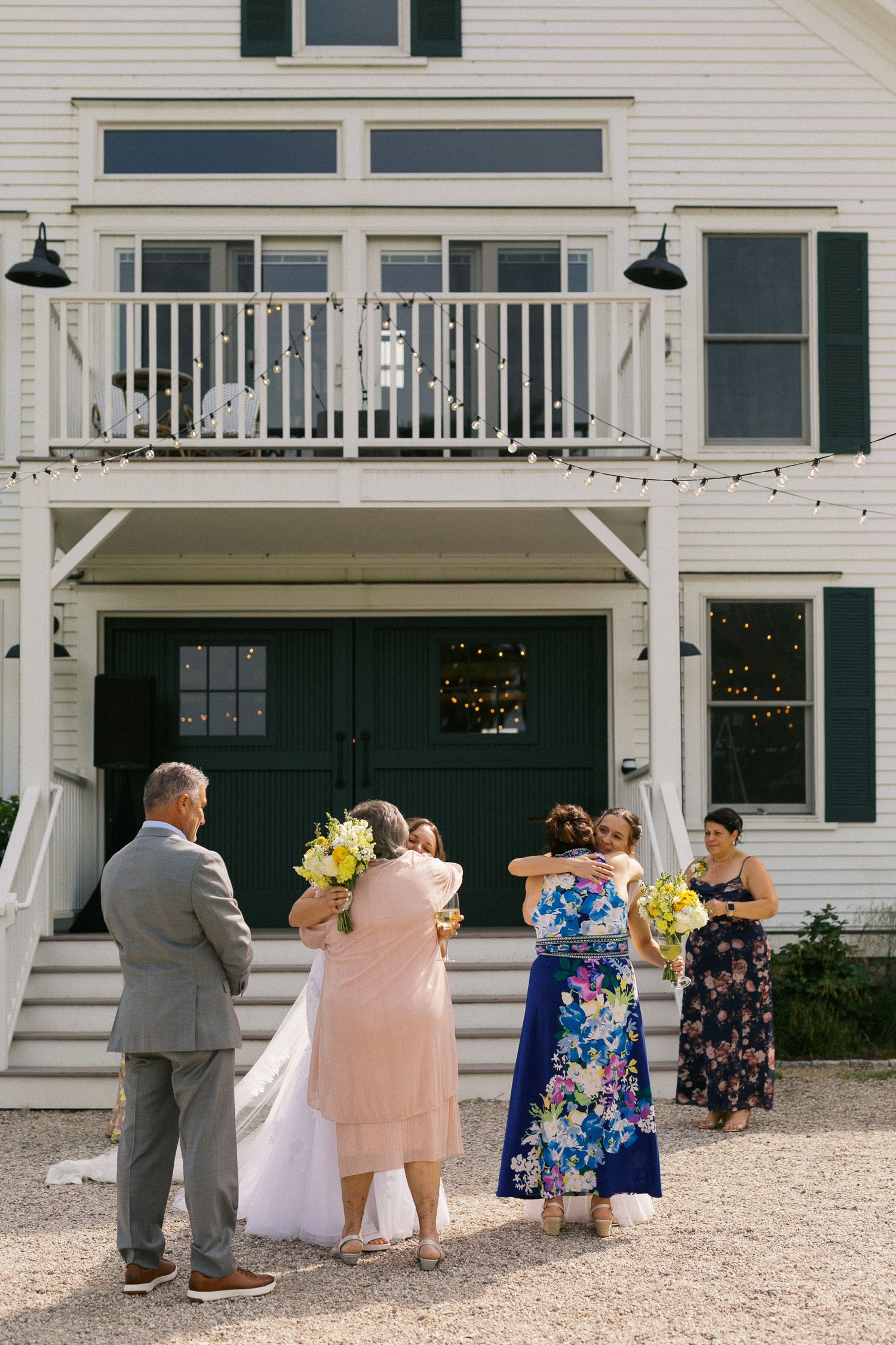 Bride and wedding guests embracing outside a white New England venue with green doors and string lights.