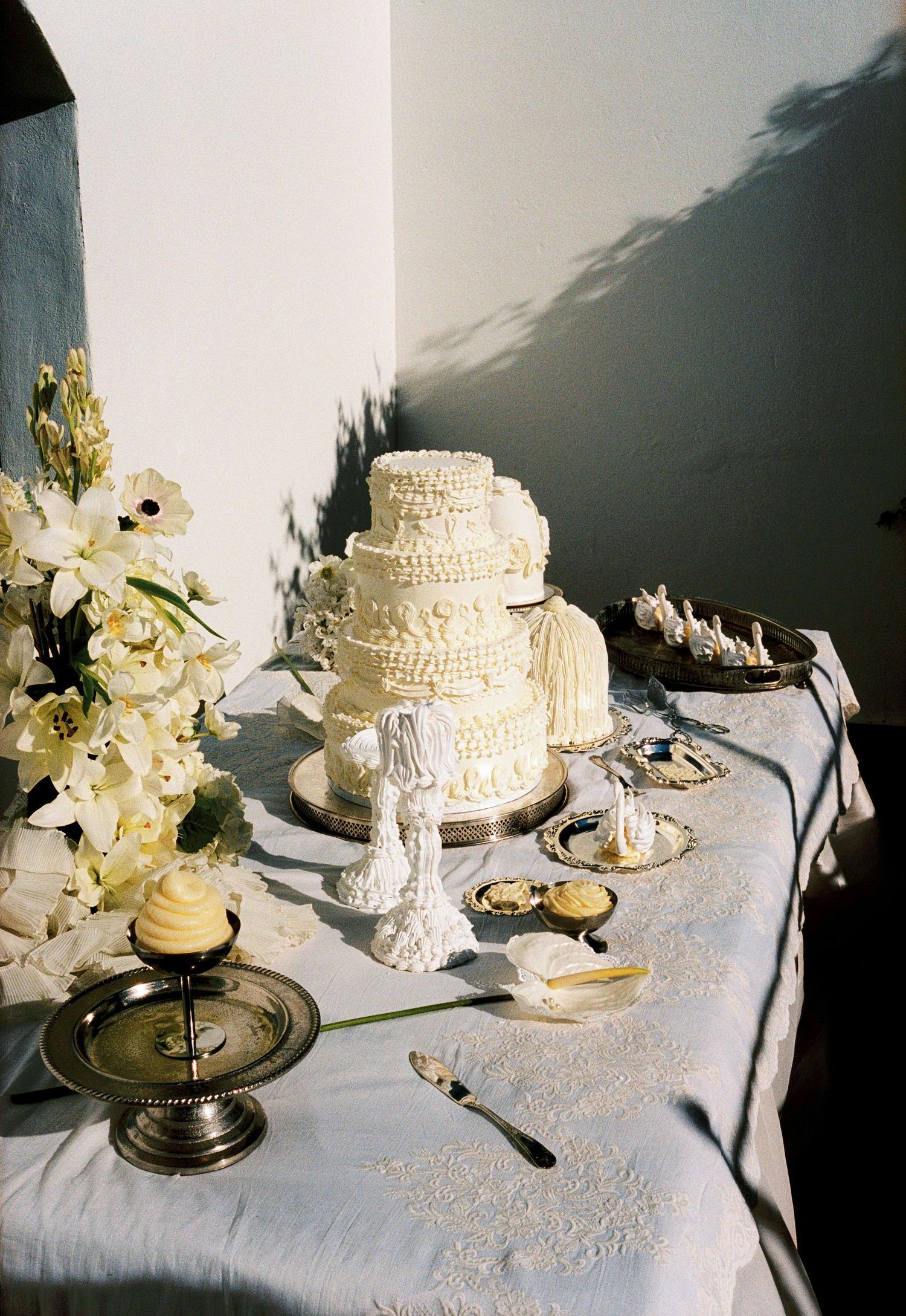 Elegant three-tier white wedding cake with ruffled frosting on a decorated table with white flowers and silver cake stands.