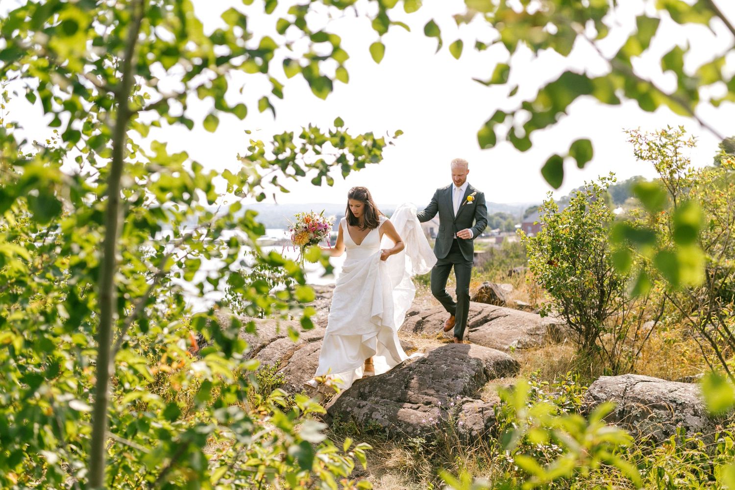 Wedding couple stands on a scenic rocky overlook surrounded by green trees during an outdoor ceremony.